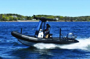 Rigid inflatable patrol boat speeds across open water, generating spray, with center console, outboard motor, radar arch, and shoreline trees under clear blue sky