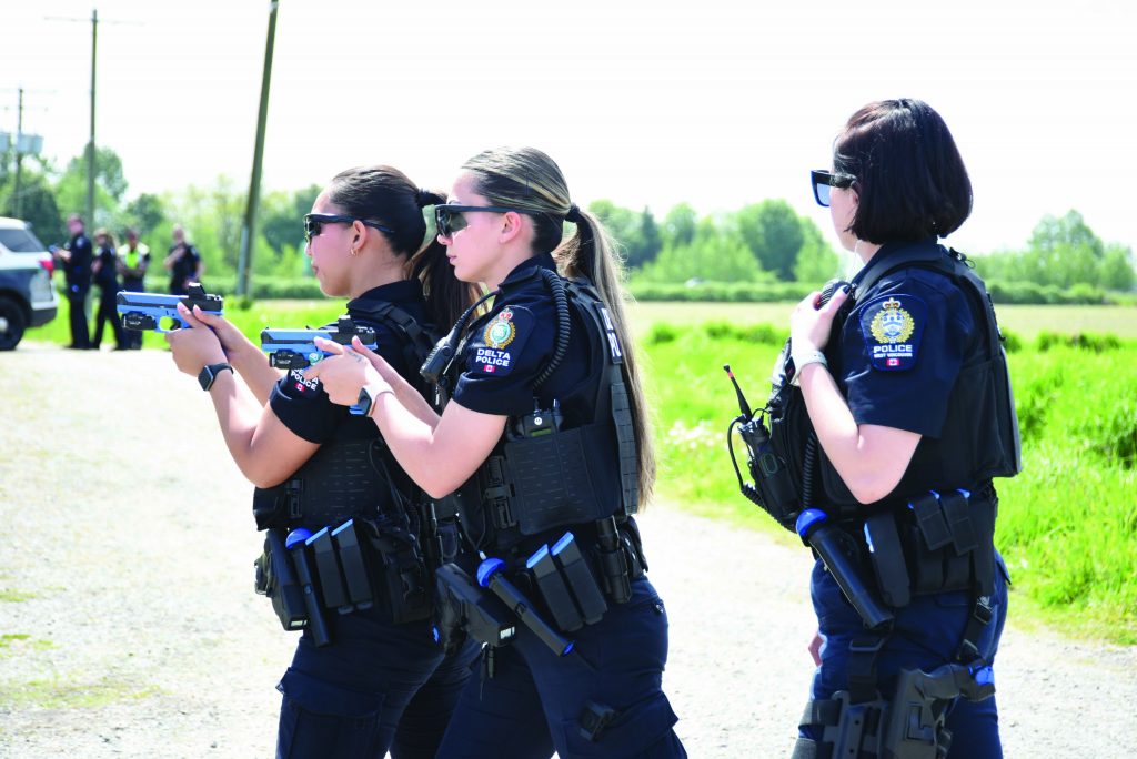 Three uniformed officers in ballistic vests and radio gear practice firearm training outdoors, aiming blue training pistols while another monitors with a handheld radio