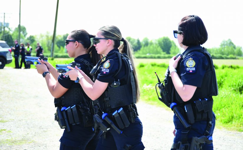 Three uniformed officers in ballistic vests and radio gear practice firearm training outdoors, aiming blue training pistols while another monitors with a handheld radio
