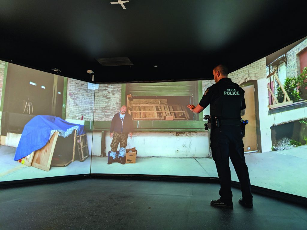 Officer stands inside a wraparound simulator projecting a warehouse scene, engaging a subject on screen while wearing duty belt, with ceiling panel and multi‑wall display