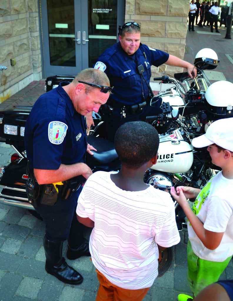 Two police officers are interacting with two children in front of a building with double glass doors. The officers are wearing dark blue uniforms and standing beside two police motorcycles. One officer is bending slightly, holding an item towards the children, while the other stands nearby. The child on the left has short hair and is wearing a white striped shirt, facing away from the camera. The child on the right is wearing a white cap and a light green shirt, holding an item in hand.