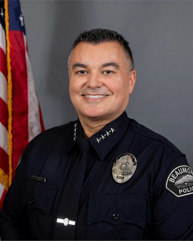 Formal studio portrait of a uniformed police executive wearing a dark dress uniform with command stars, badge, nameplate, shoulder patch, and tie, posed before a gray backdrop with a U.S. flag visible