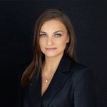 Individual with long hair wears dark blazer and small pendant necklace, standing against solid black background
