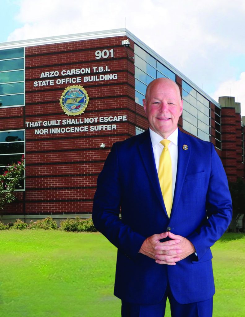 Person in navy suit with yellow tie stands in front of red brick TBI State Office Building with seal, motto, and address number 901 visible.