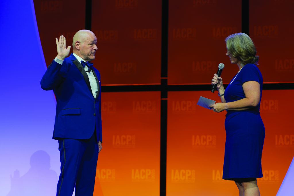 Two individuals on stage; one raises right hand in oath while the other holds a microphone and notes, with orange IACP backdrop and blue lighting