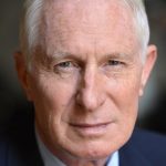 Individual wearing a dark suit jacket, white dress shirt, and patterned tie with a subtle red design, posed against a plain dark background