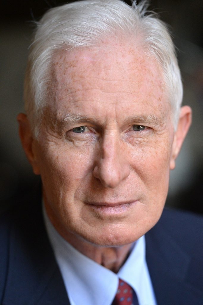 Individual wearing a dark suit jacket, white dress shirt, and patterned tie with a subtle red design, posed against a plain dark background