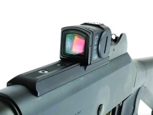 Close-up of firearm’s red dot sight mounted on top rail; illuminated reticle visible through lens with adjustment buttons on side of optic housing