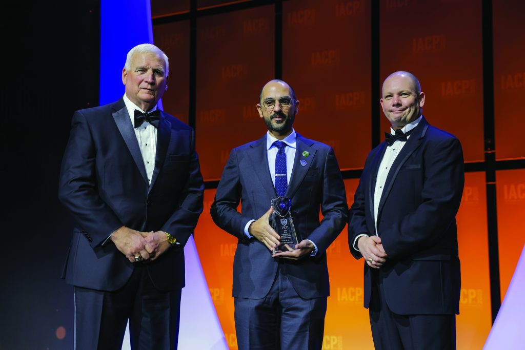 Three individuals in formal suits stand on stage with orange IACP backdrop; center person holds a glass award while others pose under bright event lighting.