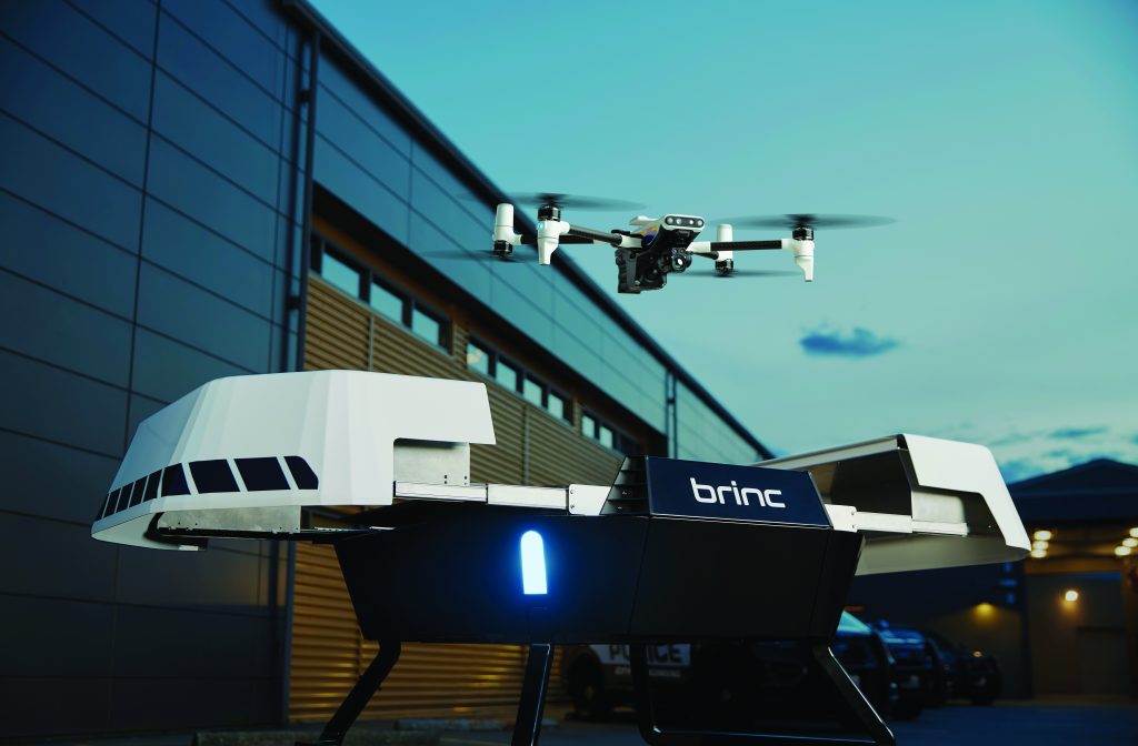 A drone responder flying away from a station, carrying equipment. The scene is set in front of a modern building with large windows, and the responder is captured mid-stride, emphasizing the urgency and readiness of their role.
