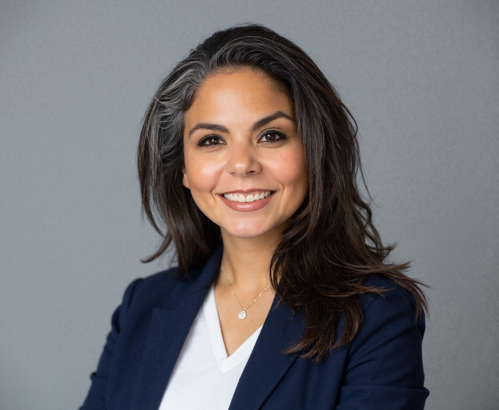 Person wearing a navy blazer over a white V-neck top with a delicate pendant necklace, standing against a plain gray background with arms partially visible