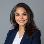 Person wearing a navy blazer over a white V-neck top with a delicate pendant necklace, standing against a plain gray background with arms partially visible