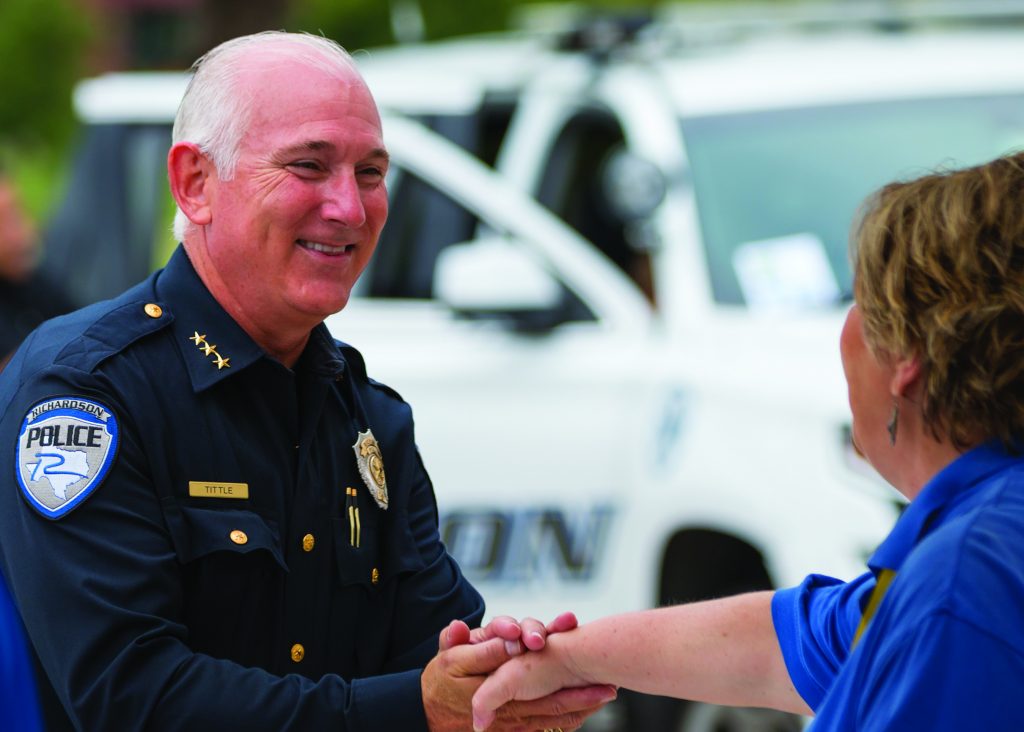 A police officers from Richardson Police Department standing together in front of their car. They are dressed in formal uniforms, showcasing leadership and camaraderie within the department shaking hands with a civilian