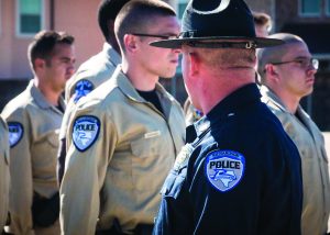 A group of law enforcement officers stands in formation outdoors. The officers are dressed in beige uniforms with blue patches on their sleeves that read "POLICE" and feature an emblem. In the foreground, a senior officer wearing a dark blue uniform and a wide-brimmed hat is facing the group, with his back to the camera. The senior officer's uniform also has a similar "POLICE" patch on the sleeve, along with additional insignia and badges indicating rank or department affiliation. The background shows part of a building with windows, suggesting an outdoor training or briefing session.