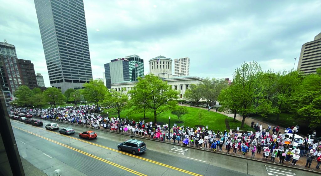 Large protest crowd lines sidewalk along a city street beside a government building and park, holding signs, with vehicles passing and tall office towers under cloudy sky