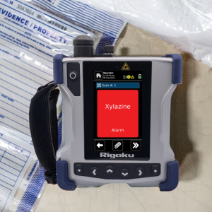 Rigaku handheld analyzer with red 'Xylazine Alarm' alert screen; background shows evidence bags labeled 'EVIDENCE/PROPERTY' and white substance