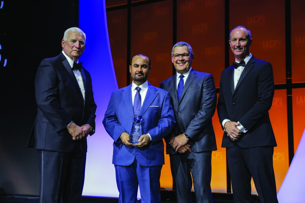 Four individuals in formal suits stand on stage with orange IACP backdrop; center person holds a glass award while others pose under bright event lighting.