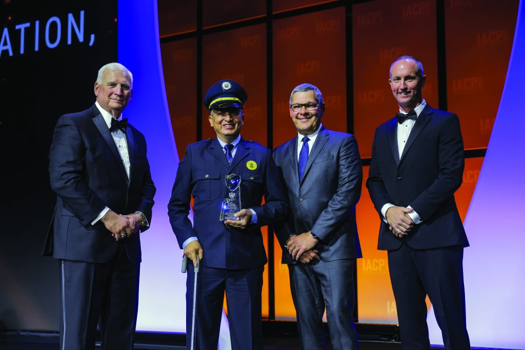 Four individuals in formal attire stand on stage with orange and purple backdrop; one in police uniform holds a glass award and a cane