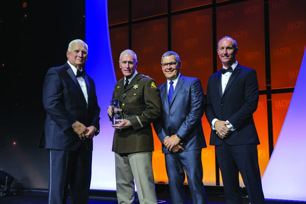 Four individuals in formal attire stand on stage with orange and purple backdrop; one in brown sheriff uniform holds a glass award