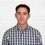 Individual in black and white checkered button-up shirt stands before white brick wall