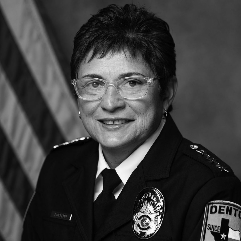 A black and white photograph of a police officer in uniform. The uniform includes a dark jacket with several buttons, epaulets on the shoulders, and various insignia. On the left side of the chest, there is a name tag that reads "Catherine." Below the name tag is a badge with intricate detailing. On the right shoulder, there is an embroidered patch that reads "Denton Police" with an outline of Texas and the word "Since" followed by a year that is not fully visible. The officer wears a tie and has short hair. In the background, there is an American flag partially visible on the left side.