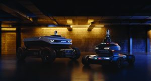 Two wheeled security robots with low-profile bodies, sensor towers, and glowing lights stand in a dim industrial space with concrete walls and overhead fixtures