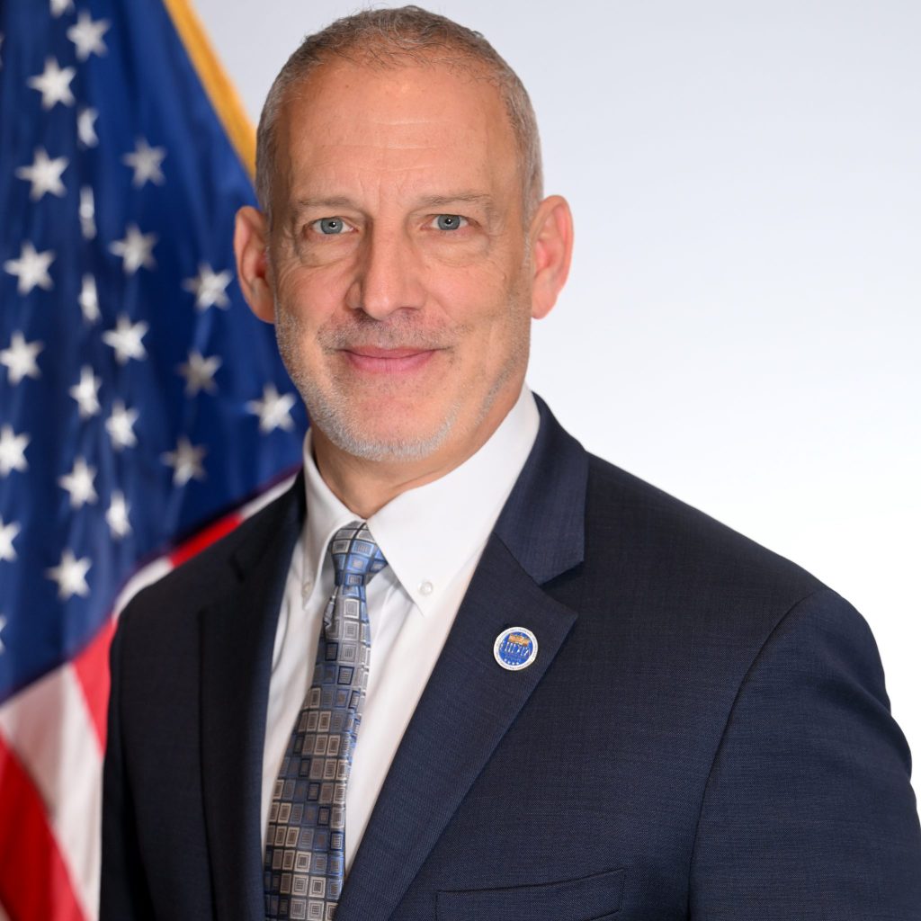 Individual in dark suit, white shirt, patterned tie, and official lapel pin stands before American flag—formal attire suggesting official capacity