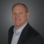 Person in dark suit jacket over light blue dress shirt stands against plain gray background