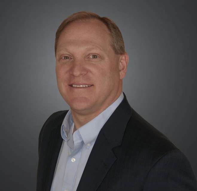 Person in dark suit jacket over light blue dress shirt stands against plain gray background