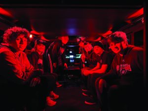 Seven people in casual attire sit inside dimly lit vehicle with red lighting, surrounding central electronic equipment and monitors