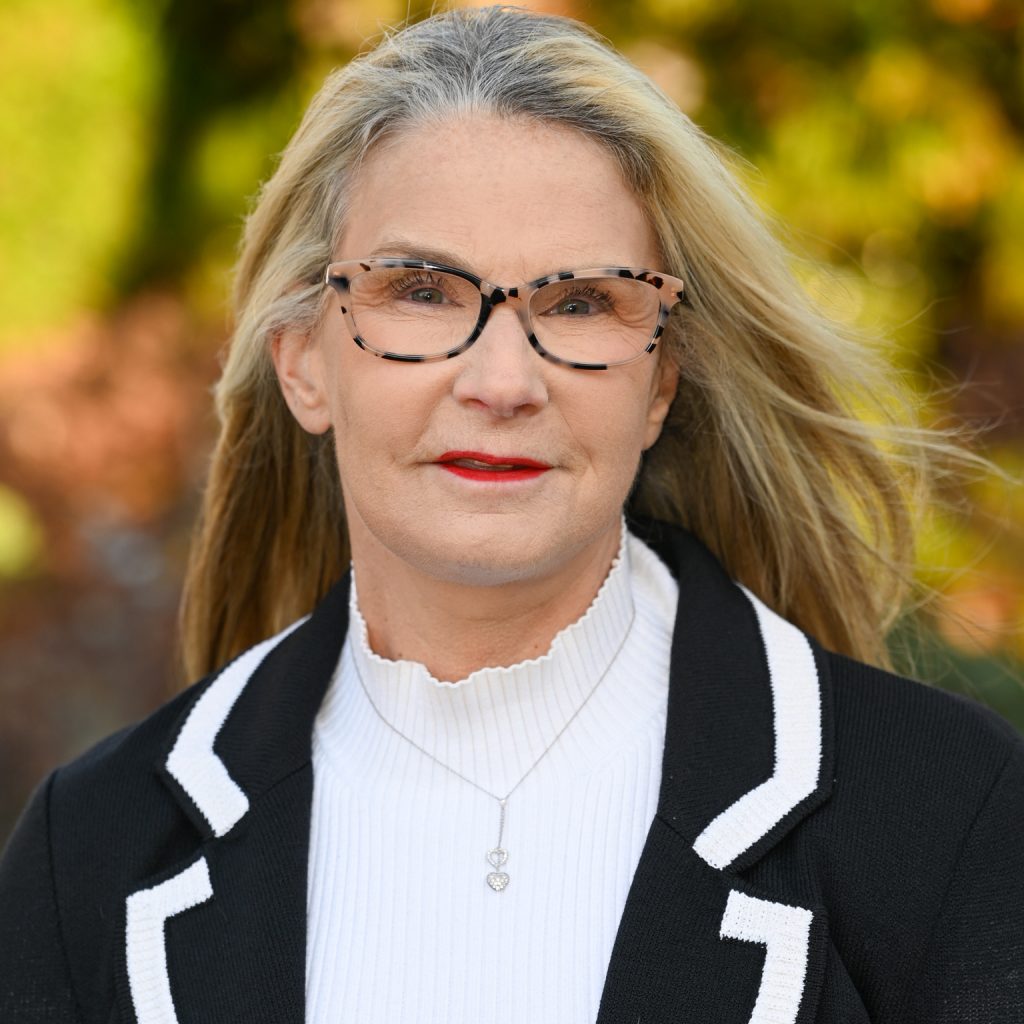 a person with long, blonde hair standing outdoors against a blurred background of green and brown foliage. The person is wearing a black blazer with white trim over a white turtleneck sweater. The person has a silver necklace with a small pendant around the neck.