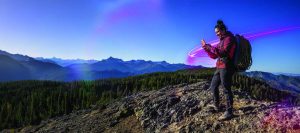 Person wearing a red jacket and large hiking backpack stands on rocky mountain peak using a smartphone, with forested slopes and distant blue mountains under clear sky