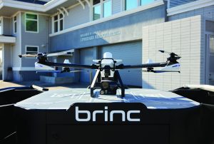 Quad‑rotor drone rests on a BRINC dock station outside a lifeguard headquarters building, showing camera module, landing pad graphics, garage doors and signage
