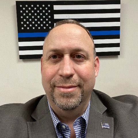 Individual in suit with blurred face stands before black-and-white American flag with blue stripe, symbolizing law enforcement support