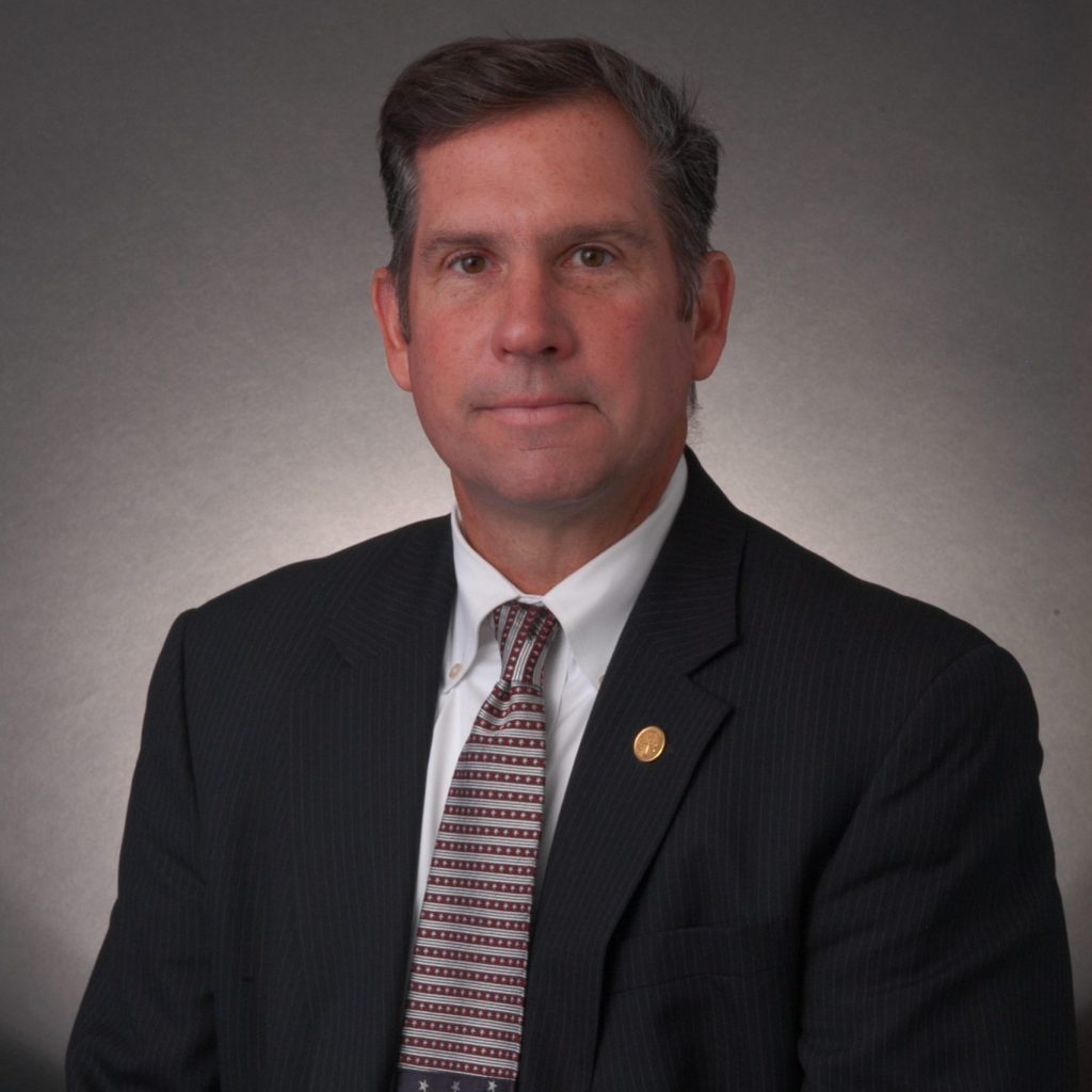 Studio portrait of an individual wearing a dark pinstripe suit, white shirt, and patterned tie with lapel pin, posed against a neutral gradient backdrop under soft lighting