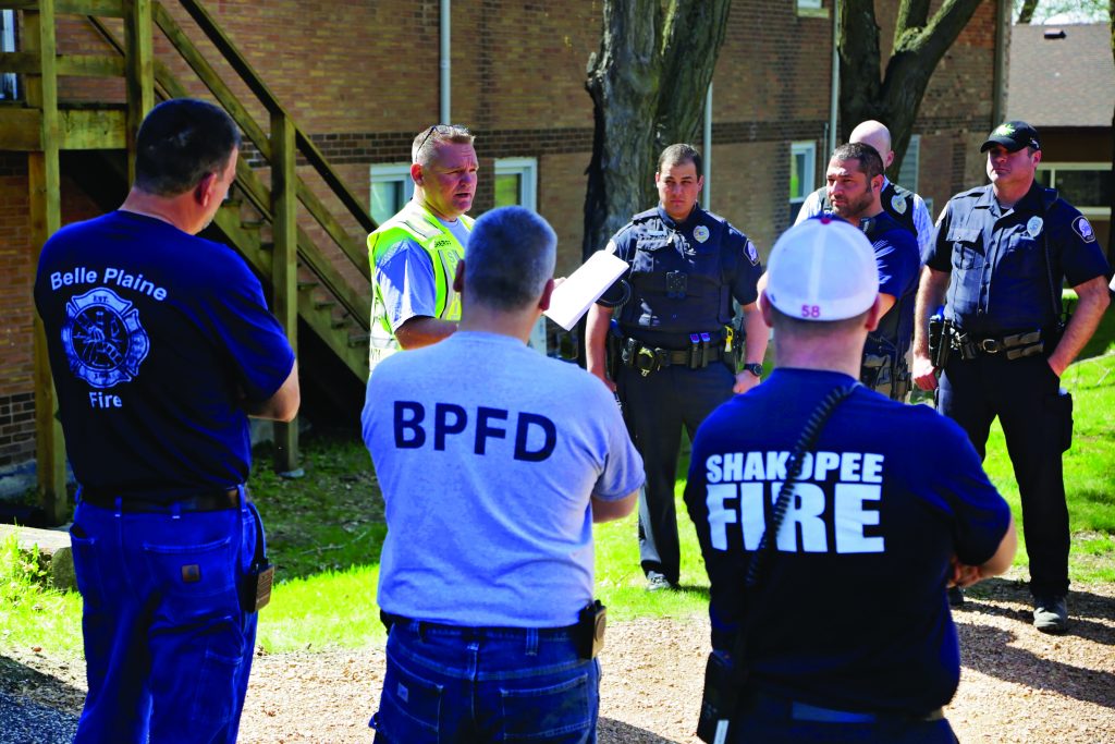 A group of eight individuals, consisting of police officers and firefighters, are gathered outdoors in a semicircle. The setting appears to be a residential area with brick buildings and trees in the background. The person at the center is holding a piece of paper and seems to be addressing the group. Three firefighters are visible from behind; one is wearing a navy blue shirt with "Belle Plaine Fire" written on it, another is wearing a light blue shirt with "BPFD" on it, and the third is wearing a navy blue shirt with "Shakopee Fire" written on it. The police officers are dressed in dark uniforms with badges and equipment belts. Some have their hands resting on their hips or crossed over their chests as they listen attentively.