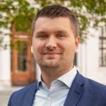 Individual in blue suit jacket and white shirt stands outdoors; background features green foliage and a building with a wooden door