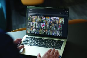 Person operates a laptop displaying a security investigation platform with searchable surveillance results, thumbnail image gallery, filters, timestamps, sorting controls, keyboard, trackpad, tabletop, chairs, and a blurred indoor workspace visible in the background