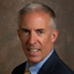 a professional portrait of an individual. The person is wearing a dark suit jacket, a light blue dress shirt, and a yellow striped tie. There is a small circular pin on the left lapel of the suit jacket. The background is plain and brownish in color, giving the image a formal appearance.