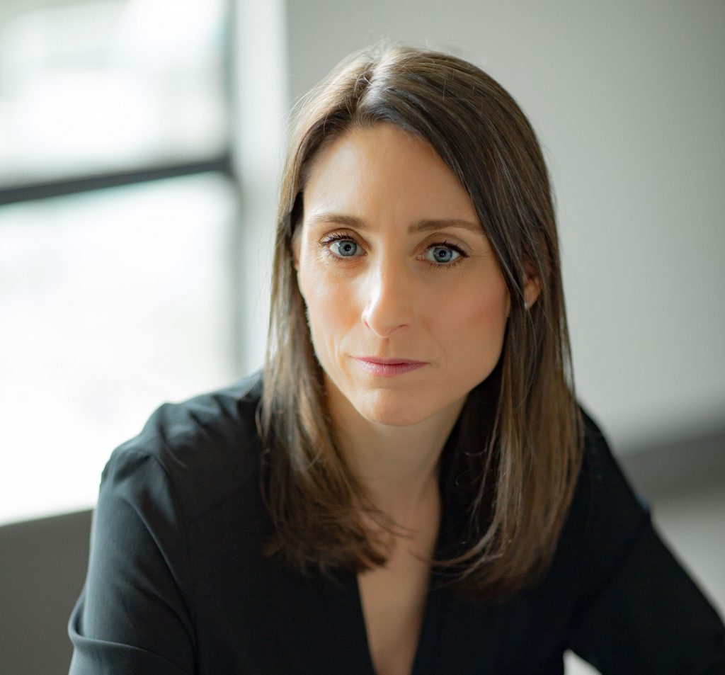 Person with straight shoulder-length brown hair wearing a black V-neck blouse, seated indoors near a window with soft natural light and neutral background