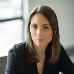 Person with straight shoulder-length brown hair wearing a black V-neck blouse, seated indoors near a window with soft natural light and neutral background