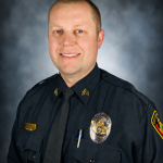 Uniformed police sergeant in dark dress uniform with tie, badge, nameplate, collar chevrons, shoulder patch, pen, and buttoned pockets, posed against a blue-gray studio backdrop with professional portrait lighting and a chest-up composition