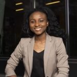 Person in beige blazer and black top stands confidently before a building, wearing hoop earrings and a necklace; face intentionally blurred