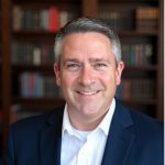 Person wearing a dark navy suit jacket over a white collared shirt, standing in front of a wooden bookshelf filled with books of various sizes and colors.