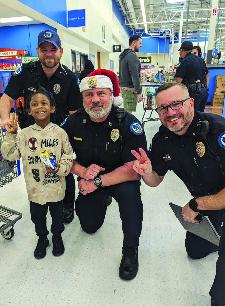 Three uniformed officers kneel and stand beside a child holding snacks in a store aisle; one officer wears a Santa hat, shopping carts visible in background