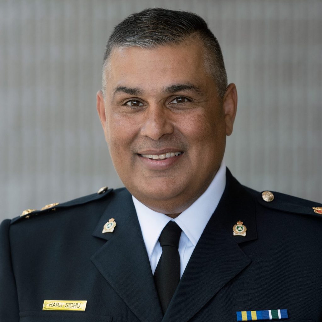 Uniformed officer in dark dress uniform with gold buttons, collar insignia, ribbon bars, and nameplate reading HARJ SIDHU stands before a neutral backdrop
