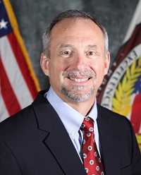 Person in a dark suit jacket, light blue dress shirt, and red patterned tie standing before U.S. flag and official seal on a textured gray backdrop