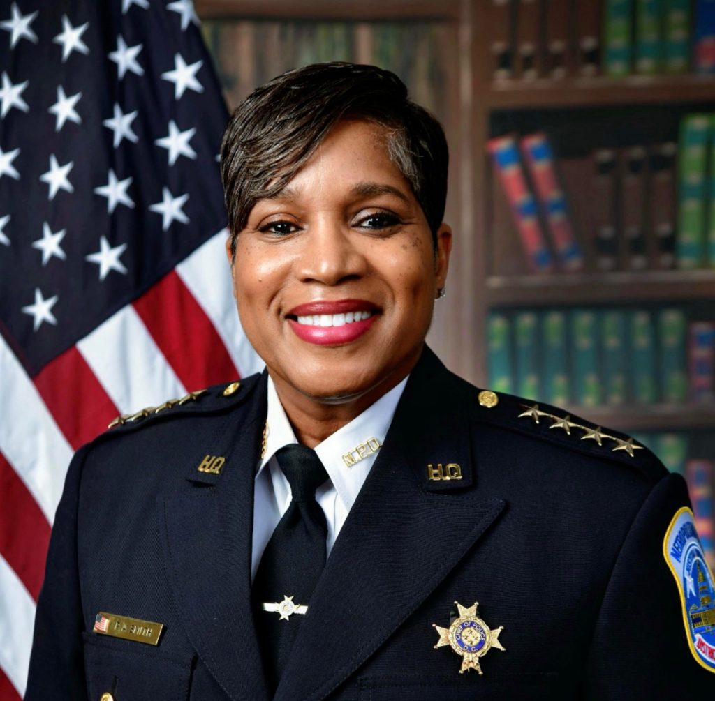 Person wearing a dark formal law enforcement uniform with gold buttons, shoulder stars, nameplate, and badge; U.S. flag and bookshelf in background