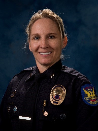 Police officer in dark uniform with gold badge, collar pins, and Phoenix Arizona patch on sleeve, standing against a dark blue studio backdrop
