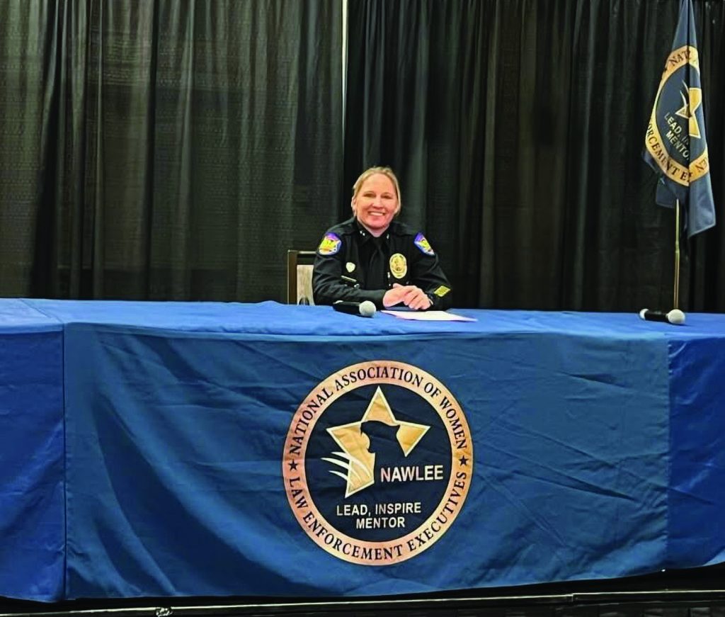 Uniformed officer seated at a table with NAWLEE logo and slogan ‘Lead, Inspire, Mentor’; two microphones on table, flags and black curtain backdrop